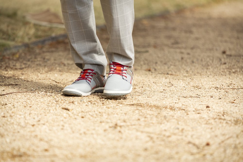 Eine Person in ZAQQs QANBERRA Grauer Hose und hellgrauen Schuhen mit roten Schnürsenkeln, möglicherweise Barfußschuhe aus Nappaleder, steht auf einem Feldweg. Der Hintergrund ist mit Gras und unscharfer Umgebung gefüllt.