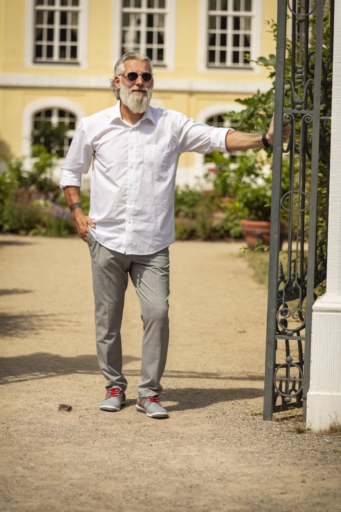 Ein Mann mit Bart und Sonnenbrille steht draußen vor einem verzierten Tor. Er trägt eine graue QANBERRA-Hose von ZAQQ und ein frisches weißes Hemd. Sein Look wird durch Freizeitschuhe aus Nappaleder vervollständigt. Im Hintergrund ist ein gelbes Gebäude mit großen Fenstern von leuchtendem Grün umgeben.