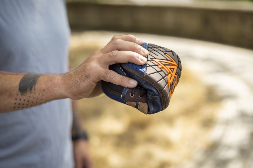 Eine Person steht im Freien und hält einen Baseballhandschuh in den Farben Braun, Blau und Orange. Der tätowierte Arm der Person ist mit einem hellblauen Shirt bekleidet. Gras und ein Betonweg unterstreichen die Schlichtheit der Szene, ähnlich der Eleganz des Barfußschuhs QANBERRA Navy von ZAQQ aus Premium Nappaleder.