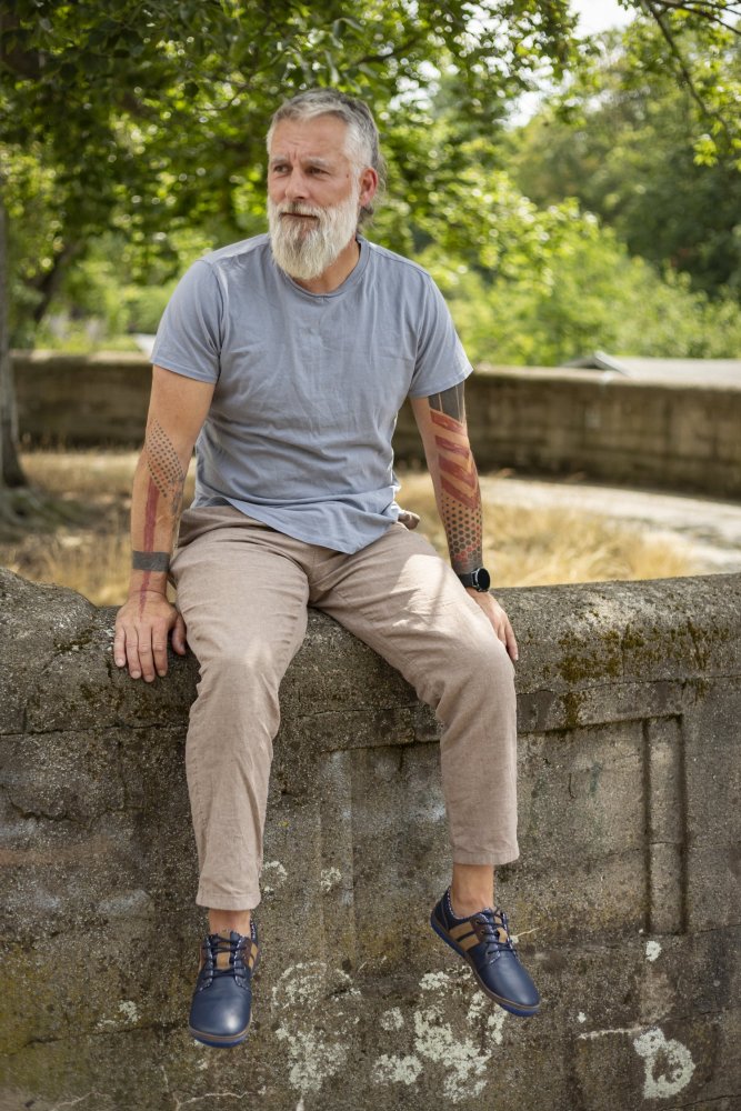 Ein bärtiger Mann mit Tattoos sitzt auf einem Steinvorsprung in einem Park und präsentiert seine ZAQQ QANBERRA Navy Schuhe mit minimaler Sohle. Er trägt ein hellblaues T-Shirt, beige Hosen und das blaue Schuhwerk aus hochwertigem Nappaleder. Im Hintergrund blühen Bäume und Grünpflanzen.