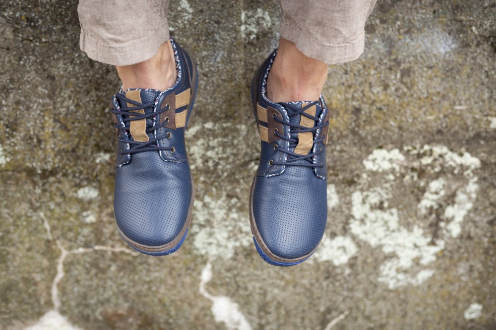 Eine Person trägt QANBERRA Navy-Schuhe von ZAQQ, aus blauem Leder mit braunen Akzenten und perforierten Details, kombiniert sie mit beigen Hosen, während sie auf einer strukturierten Steinoberfläche steht. Diese hochwertigen Schuhe aus Nappaleder haben Schnürsenkel, die dem lässigen Outfit eine stilvolle Note verleihen.