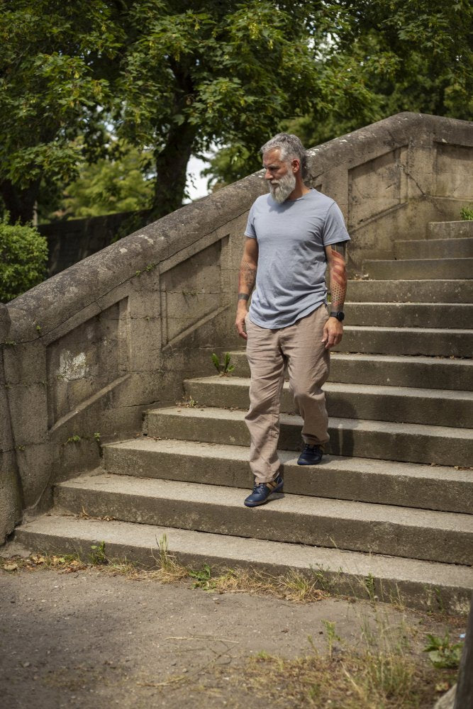 Ein Mann mit grauem Bart und Tattoos läuft Betonstufen in einem Park hinunter. Er trägt ein hellgraues T-Shirt, beige Hosen und ZAQQs QANBERRA Navy Barfußschuhe mit minimaler Sohle. Im Hintergrund sind grüne Bäume und Sträucher zu sehen.