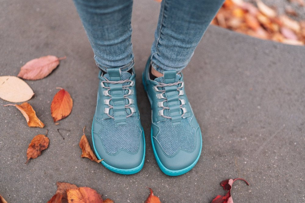 Eine Person, die die QARLY Turquoise Sneakers von ZAQQ trägt, kombiniert mit Jeans, steht auf einer Betonfläche, umgeben von verstreuten Herbstblättern in Orange- und Rottönen.