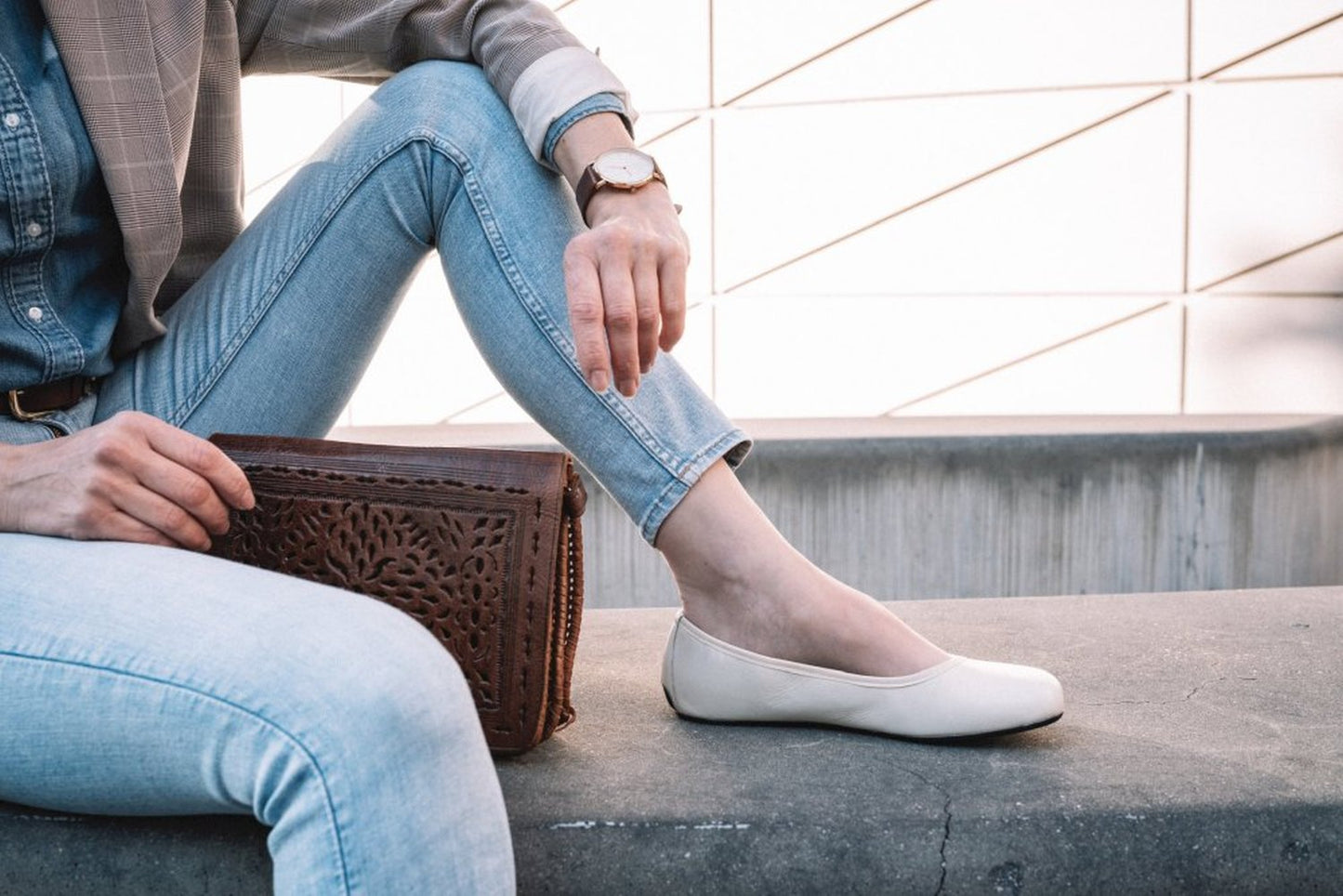 Eine Person sitzt auf einer Betonbank. Sie trägt hellblaue Jeans und ein Jeanshemd unter einem karierten Blazer. Sie ergänzt ihr Outfit mit beigen QARMA-Ballerinas von ZAQQ und hält eine braune Lederhandtasche mit aufwendigen Mustern in der Hand. Eine Uhr ziert ihr Handgelenk, vor dem geometrischen Hintergrund hinter ihnen.