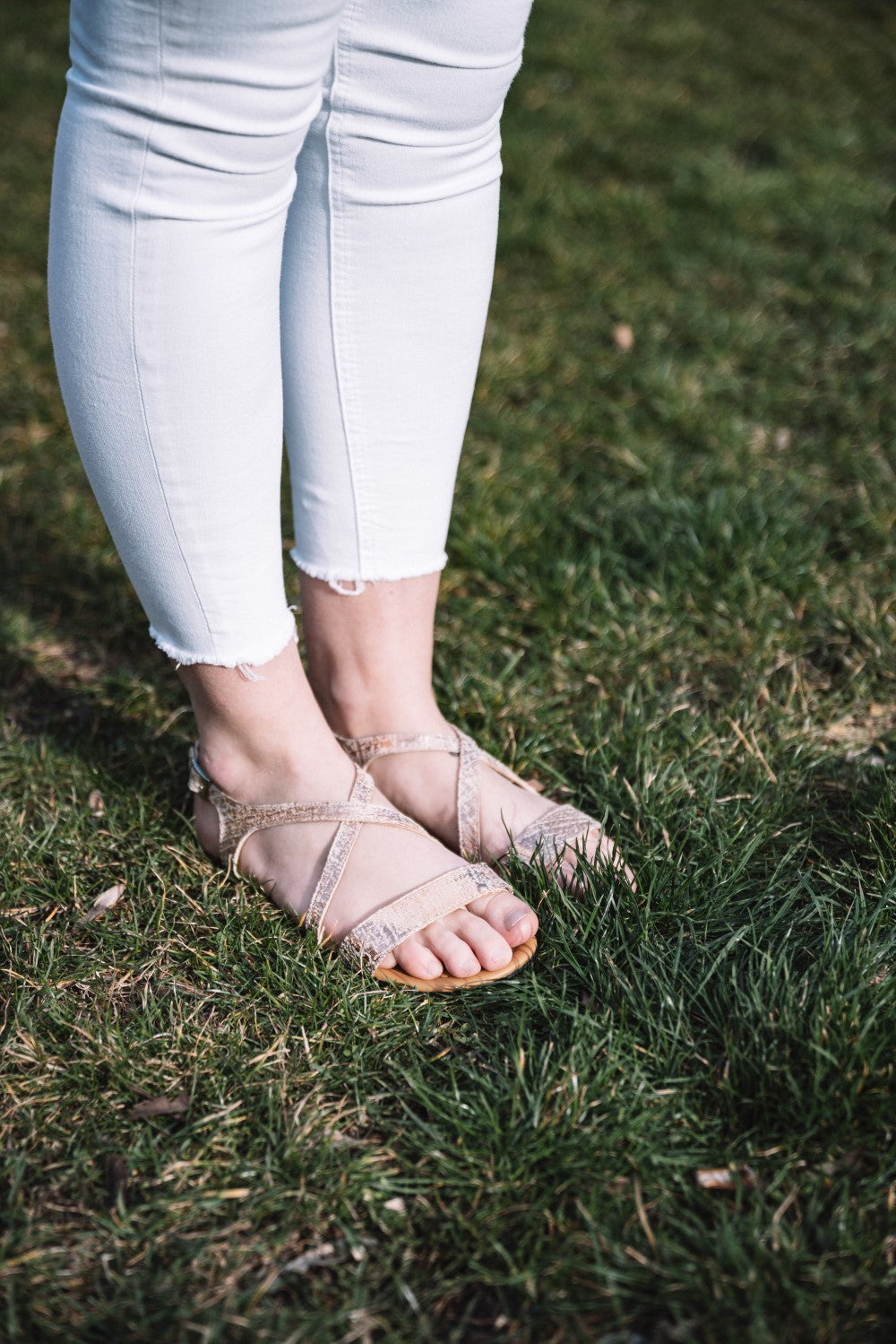 Eine Person in weißer Jeans und den Sandalen QEE-Taupe von ZAQQ mit flexibler Sohle steht auf grünem Gras. Das Sonnenlicht wirft sanfte Schatten auf den Boden und hebt das moderne Design des Schuhwerks hervor.