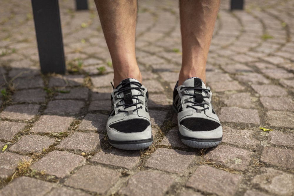 Eine Nahaufnahme einer Person, die die schwarz-grauen QENDRIQ-Sneaker von ZAQQ trägt und auf einem Kopfsteinpflasterweg steht. Die Beine sind von den Knien abwärts sichtbar, wobei ein Bein leicht hinter dem anderen liegt, was das elegante Design und den Komfort dieser Sneaker mit der MINIMAL Sohle unterstreicht.