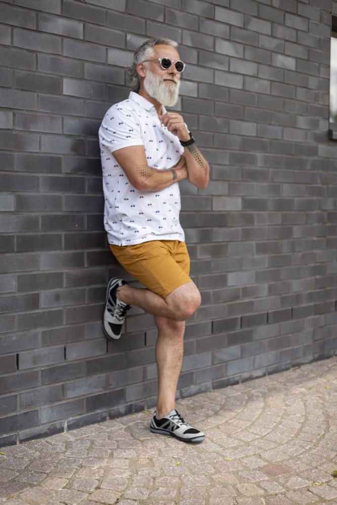 Ein Mann mit weißem Bart und Sonnenbrille lehnt an einer grauen Backsteinwand. Bekleidet mit einem gemusterten weißen Hemd, senffarbenen Shorts und schwarzen Sneakers mit dem Farbton QENDRIQ Black Grey von ZAQQ posiert er nachdenklich mit einer Hand am Kinn und einem übereinandergeschlagenen Bein.