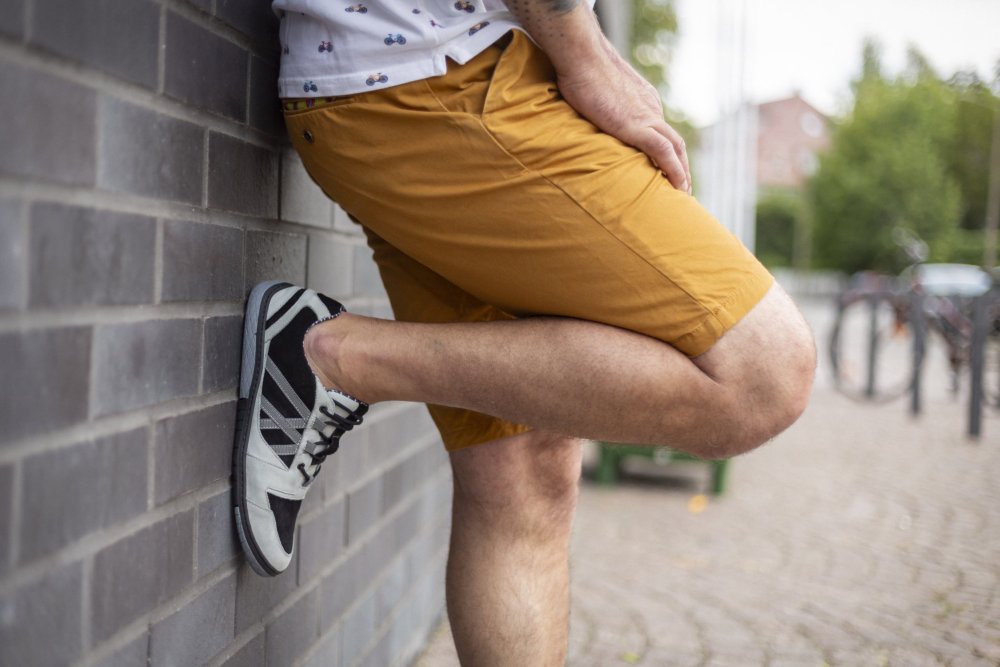 Eine Person in einem weißen Hemd mit kleinen Mustern und senffarbenen Shorts lehnt an einer Backsteinmauer und schlägt elegant ein Bein über das andere. Sie trägt die schwarz-grauen QENDRIQ-Sneakers von ZAQQ auf dem Kopfsteinpflaster, im Hintergrund parken lässig Fahrräder.