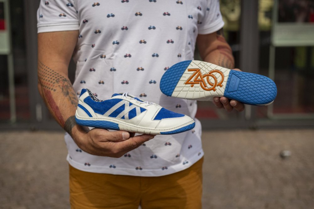 Eine Person hält ein Paar QENDRIQ Blue-Sneaker von ZAQQ. Ein Schuh steht aufrecht, während der andere ein lebendiges Sohlendesign mit den Buchstaben ZAOQ zeigt. Die Person, gekleidet in ein bedrucktes weißes Hemd, verströmt Sommerstimmung, während sie mit ihrem stylischen sommerlichen Sneaker im Freien steht.