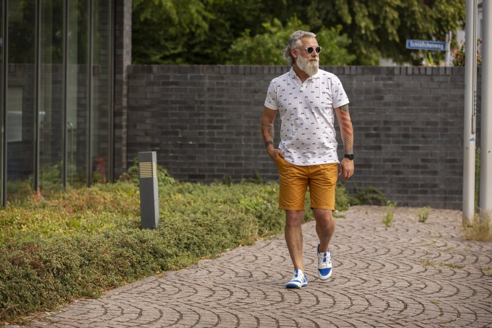 Ein Mann mit Bart und Sonnenbrille schlendert in einem weiß gemusterten Hemd, orangefarbenen Shorts und sommerlichen Sneakers „QENDRIQ Blue“ von ZAQQ auf einem gepflasterten Weg. Er steht im Freien in der Nähe eines Glasgebäudes und einer Backsteinmauer, umgeben von Grün.