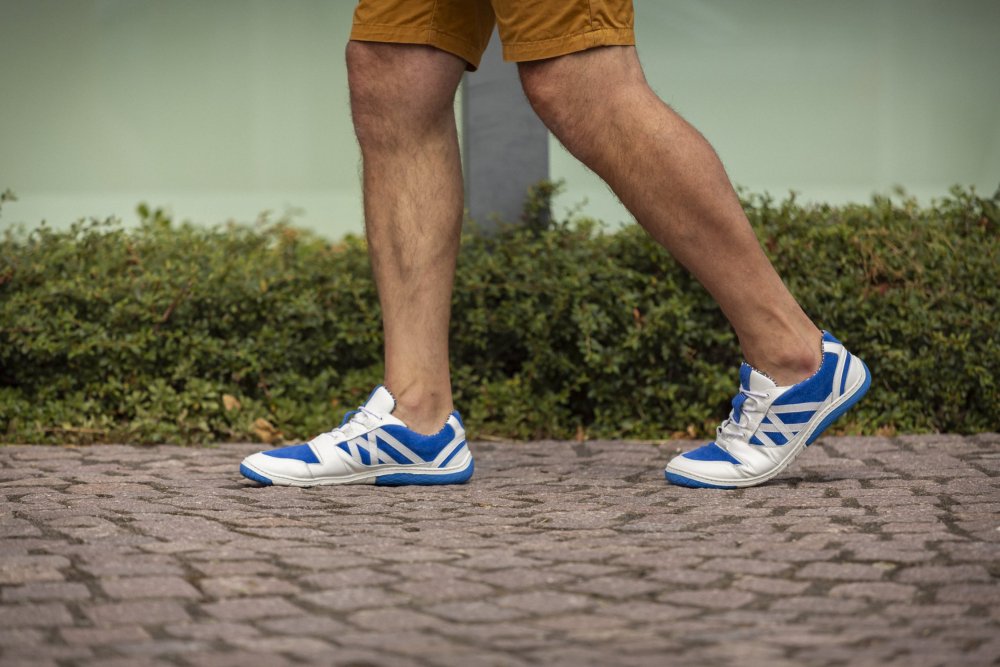 Eine Person läuft auf einem Kopfsteinpflasterweg und trägt die blauen ZAQQ QENDRIQ-Sneakers und orangefarbene Shorts. Grüne Büsche bilden eine üppige Kulisse.