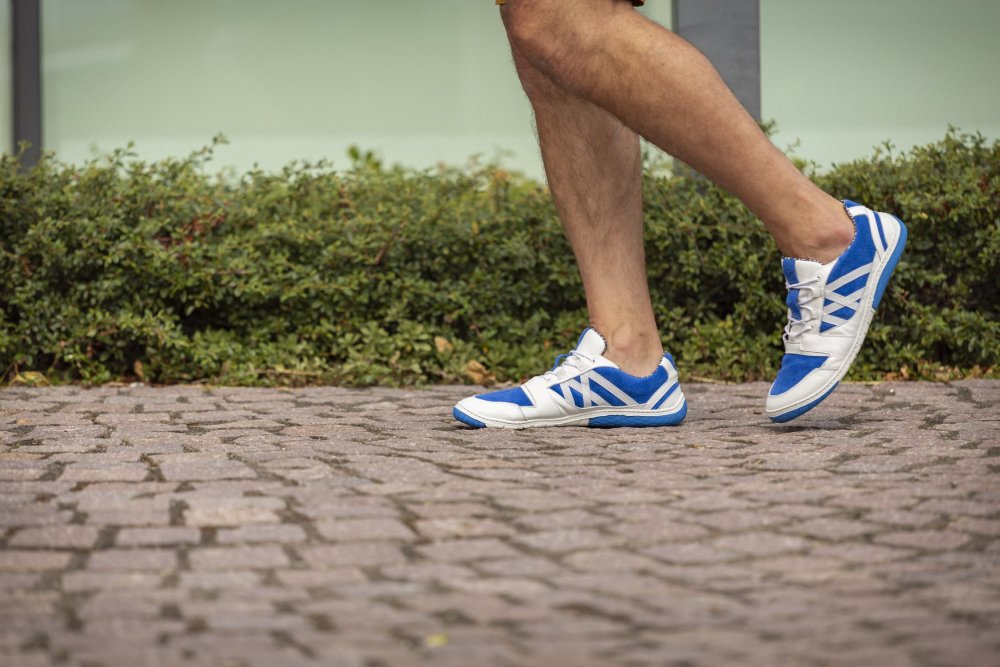 Eine Person, die die blauen QENDRIQ-Sneakers von ZAQQ trägt, läuft auf einem Kopfsteinpflasterweg, der von grünen Sträuchern gesäumt ist.