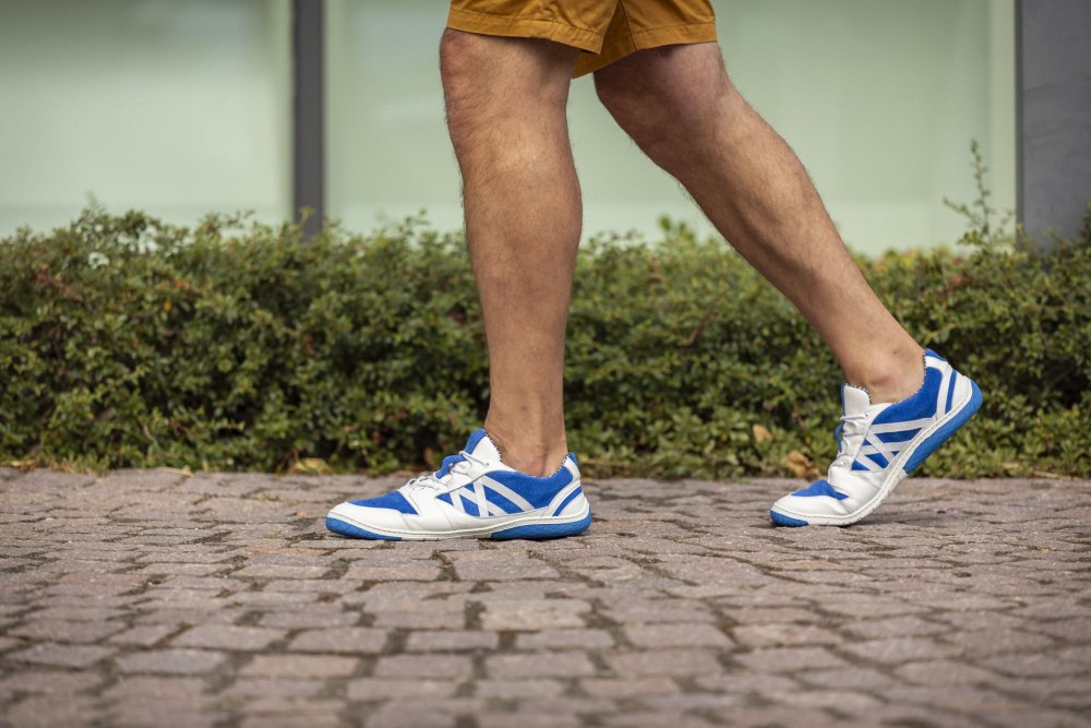 Eine Person schlendert in den blauen QENDRIQ-Sneakers und braunen Shorts von ZAQQ einen Kopfsteinpflasterweg entlang, während grünes Gebüsch eine üppige Kulisse bildet.