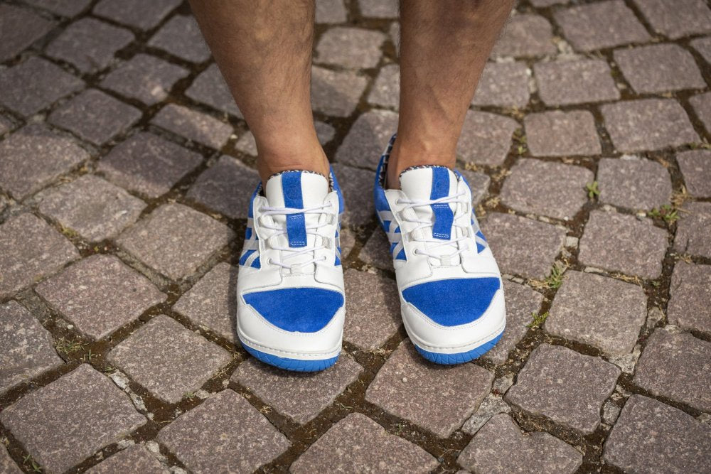 Eine Person steht auf einem Kopfsteinpflasterweg und trägt ein Paar weiße sommerliche Sneakers von ZAQQ mit deutlichen Akzenten in QENDRIQ Blue.