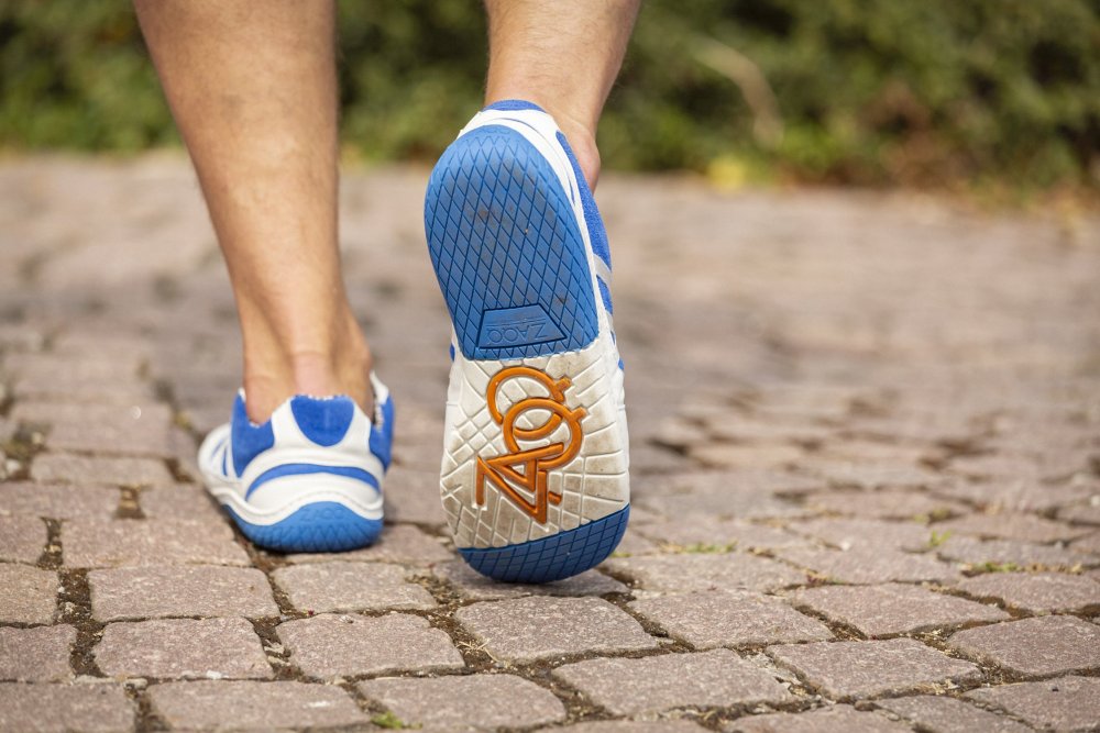 Nahaufnahme einer Person, die in sommerlichen Sneakern von ZAQQ mit blauen Akzenten auf einem Kopfsteinpflasterweg läuft. Die weißen Schuhe, die als QENDRIQ Blue-Modell entworfen wurden, zeigen auf der Sohle ein auffälliges Design mit der Nummer 404 in Orange.