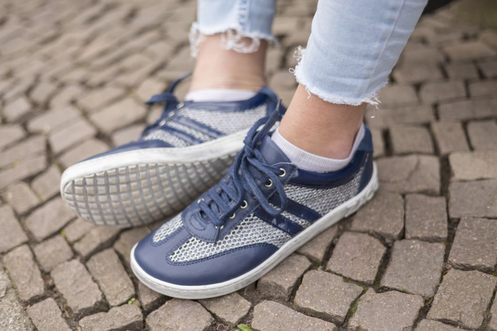 Nahaufnahme einer Person, die die blauen Barfuß-Sneakers „QIMBERLY Blue“ von ZAQQ, dazu weiße Socken und ausgefranste hellblaue Jeans trägt und auf einem Kopfsteinpflaster sitzt.