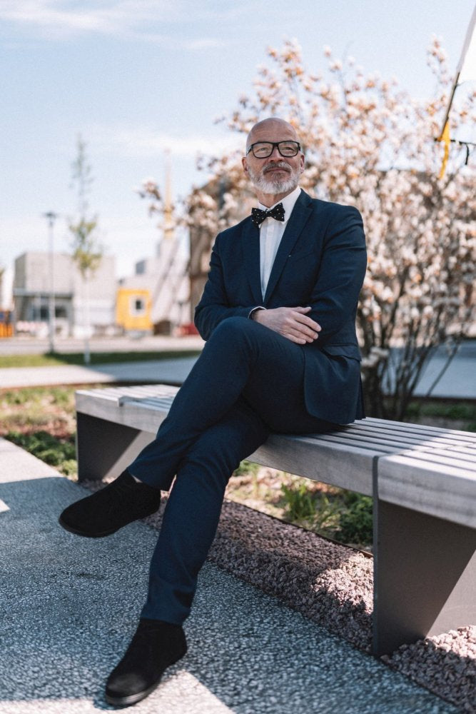 Ein Mann im Anzug, mit Brille und Fliege sitzt auf einer Bank. Er trägt stylische schwarze QLASSY-Schuhe von ZAQQ, schlägt die Beine übereinander und wirkt entspannt. Im Hintergrund zieren blühende Bäume und ein klarer Himmel den sonnigen Tag.