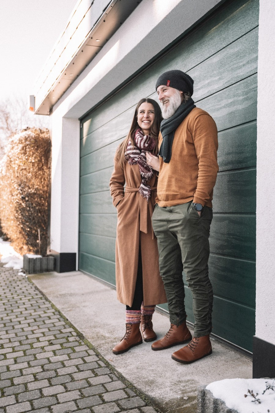 Ein lächelnder Mann und eine lächelnde Frau stehen an einem sonnigen Tag vor einer Garage. Die Frau trägt einen hellbraunen Mantel, einen gemusterten Schal und kuschelige Socken, während der Mann einen braunen Pullover, eine grüne Hose, einen Schal und eine Mütze trägt. Beide tragen die QUINTIC Winter Vegan Tabaco Stiefel von ZAQQ mit veganer Kautschuksohle. Der Boden ist teilweise mit Schnee bedeckt.