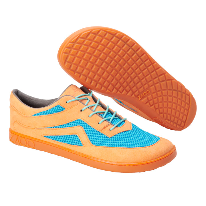 Die QLAYTON Orange Sneaker von ZAQQ bestechen durch ein blaues Mesh-Gewebe gepaart mit einer auffälligen orangefarbenen Sohle. Mit Akzenten aus Veloursleder und grauen Schnürsenkeln wird ein Schuh in der Seitenansicht präsentiert, während der andere seine einzigartige Waffelsohle hervorhebt und so die unverwechselbare Eleganz eines Barfuß-Sneakers einfängt.