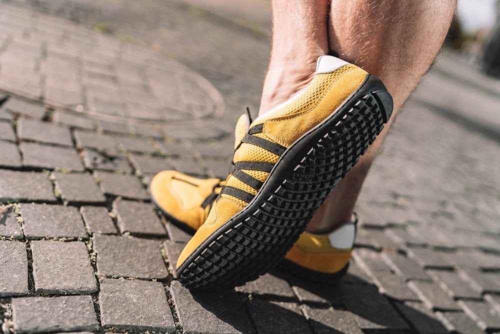 Eine Person steht auf Kopfsteinpflaster und hebt die RIQ Yellow-Sneakers von ZAQQ mit schwarzen Streifen und dicken Sohlen hervor. Diese leuchtenden Schuhe ziehen die Aufmerksamkeit auf sich und bieten ein flexibles Tragegefühl, das die strukturierte Oberfläche darunter wunderbar ergänzt.