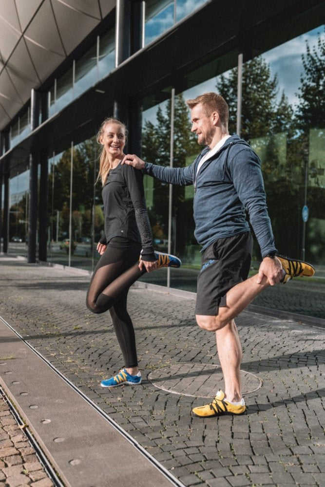 Ein Mann und eine Frau machen im Freien neben einem modernen Glasgebäude Dehnübungen. Beide tragen ZAQQ-Sportbekleidung aus dem atmungsaktiven Netzgewebe der RIQ Blue Orange-Kollektion. Lächelnd halten sie einen Fuß hinter sich, während sich Bäume in den Fenstern spiegeln.