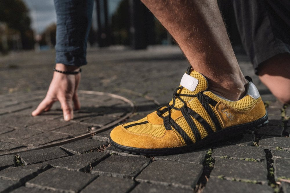 Nahaufnahme einer Person, die die RIQ Yellow-Sneakers von ZAQQ trägt und in der Startposition auf einem Kopfsteinpflaster hockt. Der Fokus liegt auf dem Schuh und hebt sein flexibles Tragegefühl hervor, wenn eine Hand den Boden berührt.