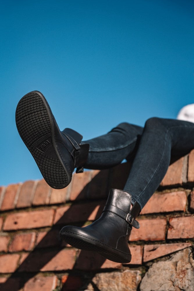 Eine Person sitzt auf einer Backsteinmauer vor einem klaren blauen Himmel und trägt Röhrenjeans und die schwarzen RIQUET-Stiefel von ZAQQ. Die dunklen Stiefel aus glattem Nappaleder baumeln über der Kante und vermitteln ein subtil stilvolles Barfußgefühl, wenn ihre Sohlen das Licht einfangen.