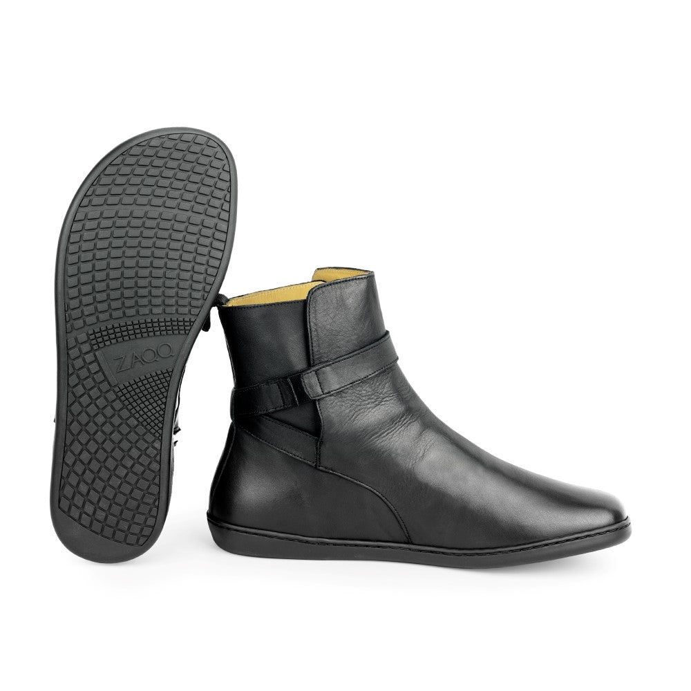 Abgebildet ist ein Paar schwarze RIQUET-Stiefeletten von ZAQQ. Ein Stiefel steht aufrecht und hebt seine glatte Lederoberfläche und das Riemchen-Boot-Detail hervor, während der andere auf der Seite liegt und die strukturierte Gummisohle und den Markennamenaufdruck zeigt.