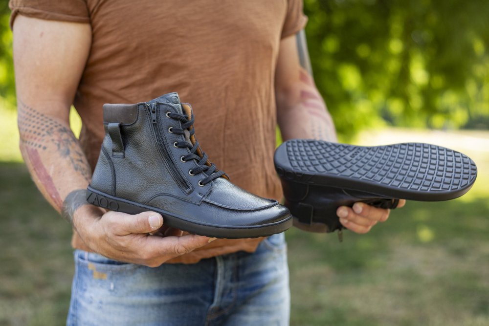 Eine Person hält in einer Hand einen eleganten ROQA Black Ankle Boot von ZAQQ und präsentiert mit der anderen die strukturierte Sohle, die den stylischen Reißverschluss hervorhebt. In einem braunen Hemd und Jeans gekleidet, steht sie im Freien vor einer Kulisse aus Gras und Bäumen.