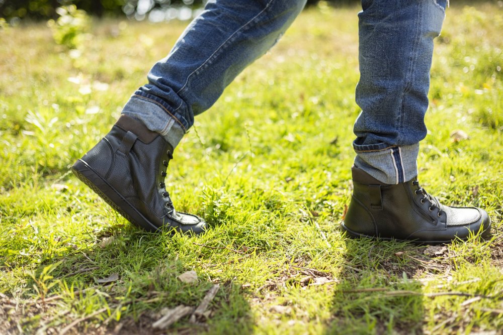 Jemand trägt hochgekrempelte Jeans und schwarze ROQA-Lederstiefel von ZAQQ mit dezentem Reißverschluss und läuft auf einem sonnenbeschienenen Grasweg.