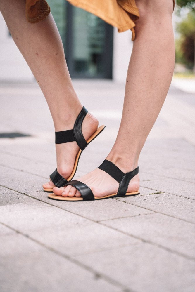 Eine Nahaufnahme von jemandem, der stylische ZAQQ SLIQ-Black-Sandalen auf einem gepflasterten Außenweg trägt. Die Sandalen haben Riemen um die Zehen und Knöchel und passen perfekt zu einem hellen Rock. Nur die Beine sind sichtbar, die den Sonnenschein an einem hellen Tag genießen.