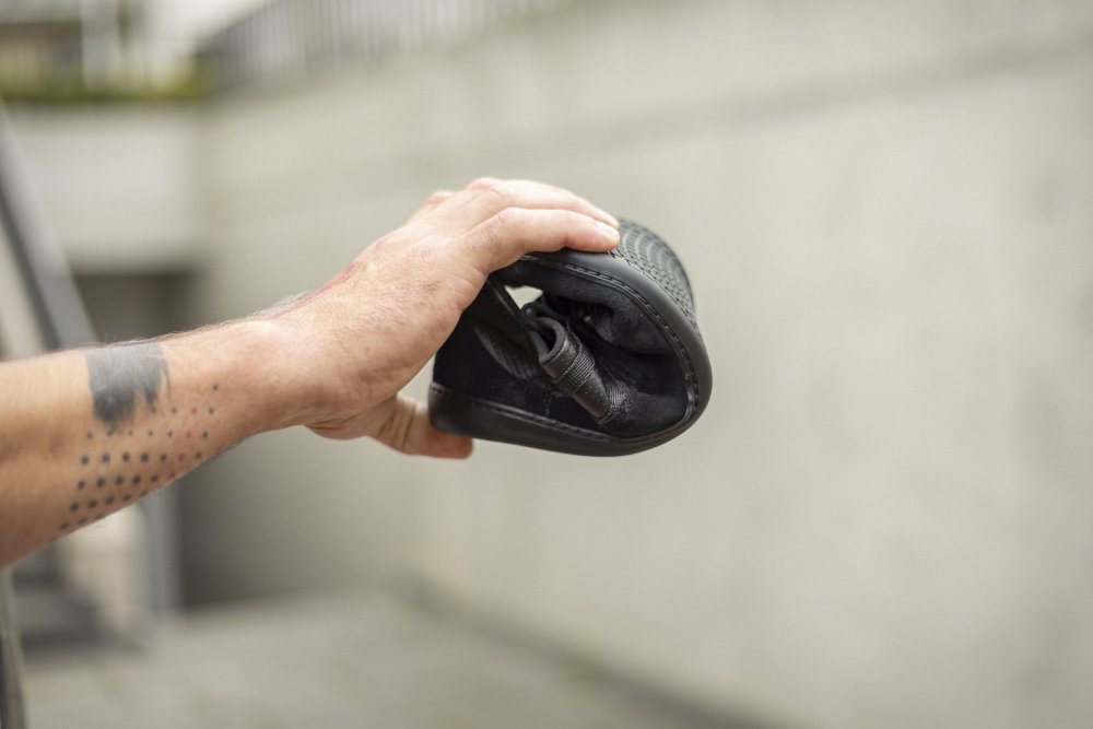 Eine Hand hält einen SPARQ Low Black von ZAQQ, der in der Mitte gefaltet ist, um seine Flexibilität hervorzuheben. Auf dem Unterarm der Person ist eine Tätowierung aus einer Reihe schwarzer Punkte zu sehen. Der unscharfe Hintergrund zeigt eine Betonoberfläche.