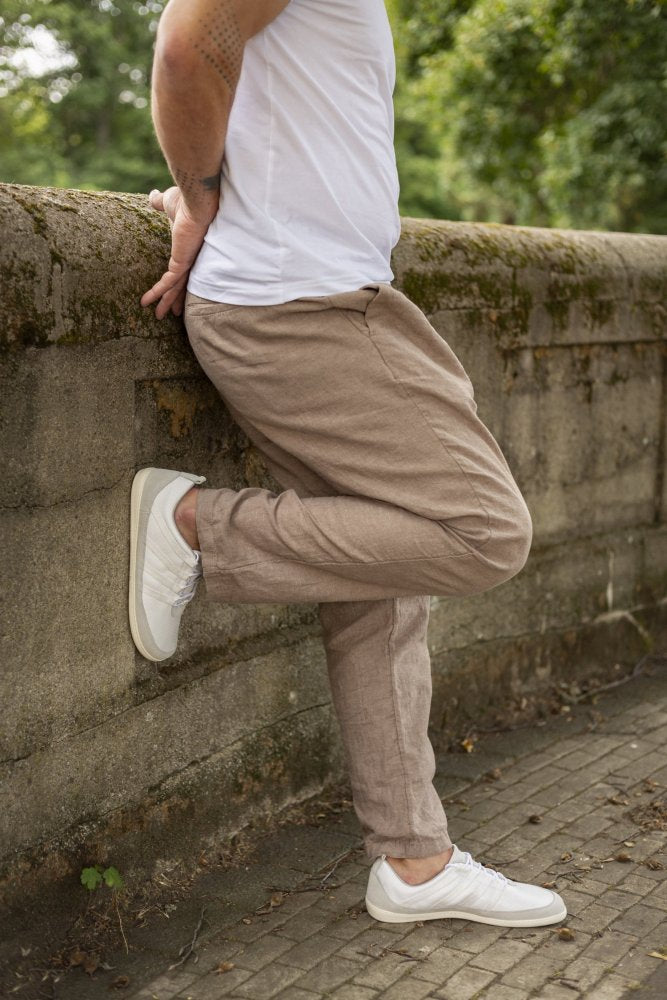 In einem weißen T-Shirt, einer beigen Hose und den SPARQ Low White Sneakers von ZAQQ lehnt eine Person im Freien an einer moosbedeckten Steinmauer und hat bequem einen Fuß über den anderen geschlagen. Im Hintergrund ist Grün zu sehen.