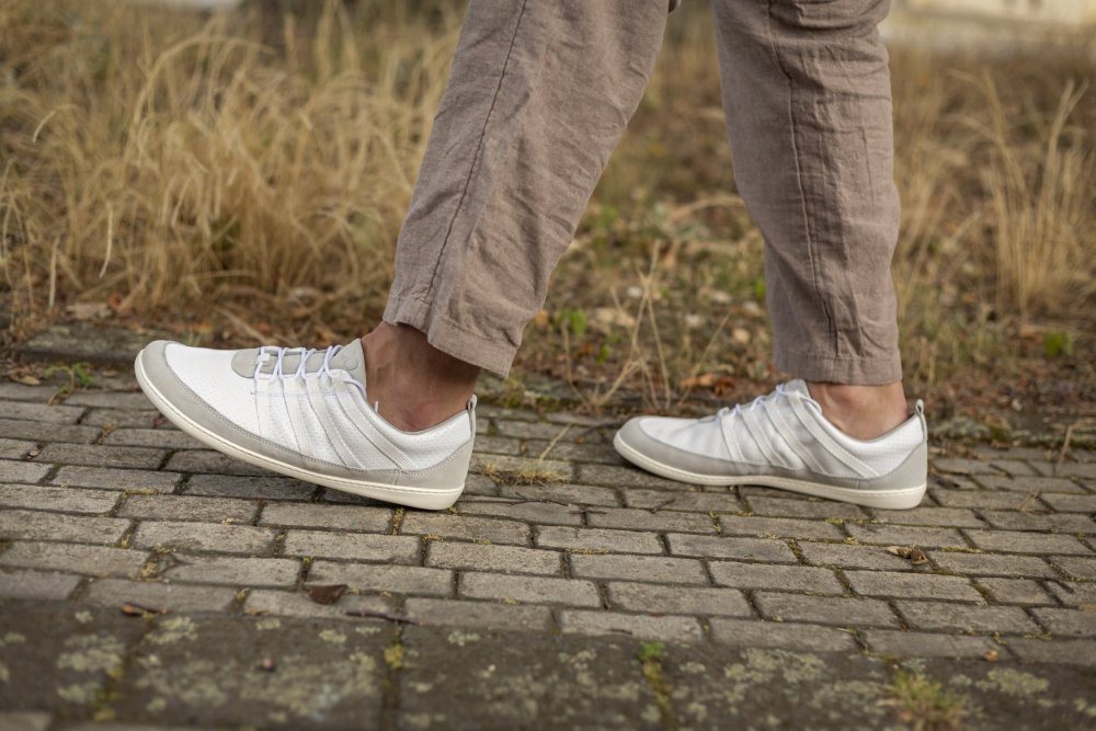 Eine Person in den SPARQ Low White Sneakers von ZAQQ läuft auf einem Kopfsteinpflasterweg mit trockenem Gras im Hintergrund. Sie trägt hellbraune Hosen, der Fokus liegt auf ihren Beinen und Schuhen, was den tadellosen Komfort bei jedem Schritt unterstreicht.