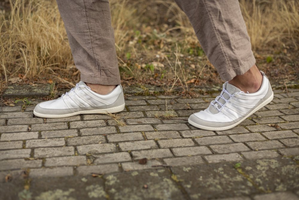 Nahaufnahme einer Person, die SPARQ Low White-Sneakers von ZAQQ und beigefarbene Hosen trägt und auf einem gepflasterten Weg geht. Der Komfort ist bei jedem Schritt spürbar. An den Rändern des Weges sind trockenes Gras und Blätter zu sehen.