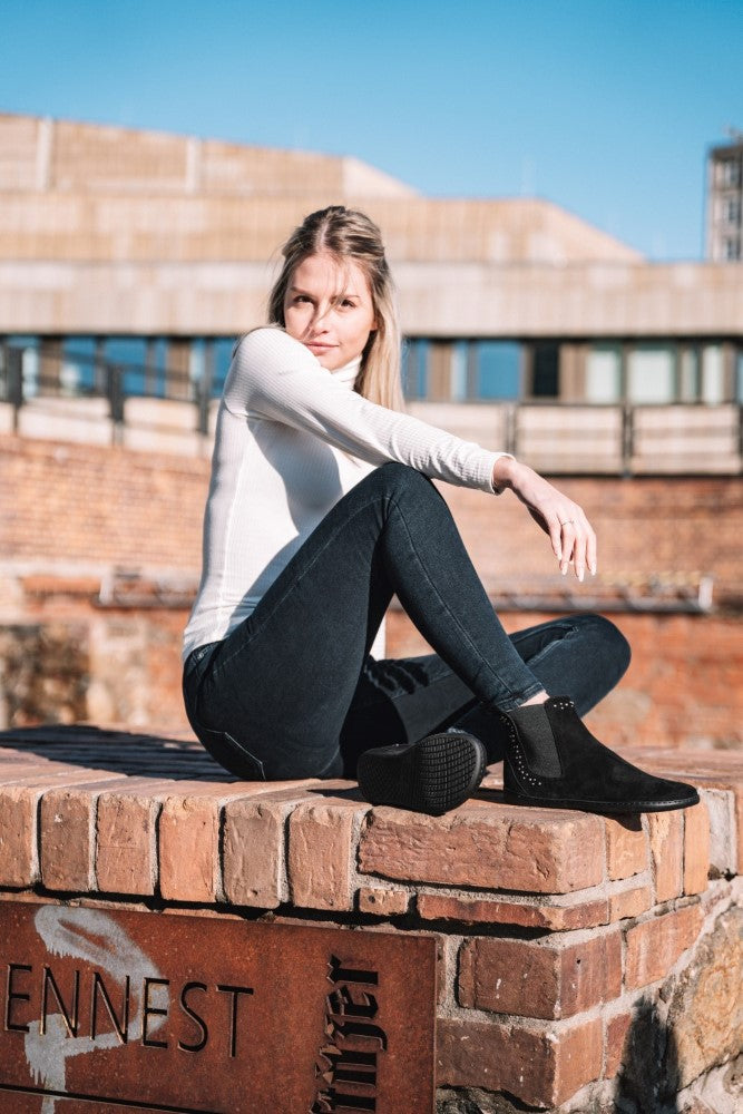 Eine Frau in einem weißen Langarmshirt und dunklen Jeans sitzt auf einer Backsteinmauer und trägt ZAQQs SPARQLE Shine Velours Black Ankle Boots, die mit dezenten Schmucksteinchen verziert sind. Sie steht im Freien vor einem verschwommenen urbanen Hintergrund unter einem klaren blauen Himmel.