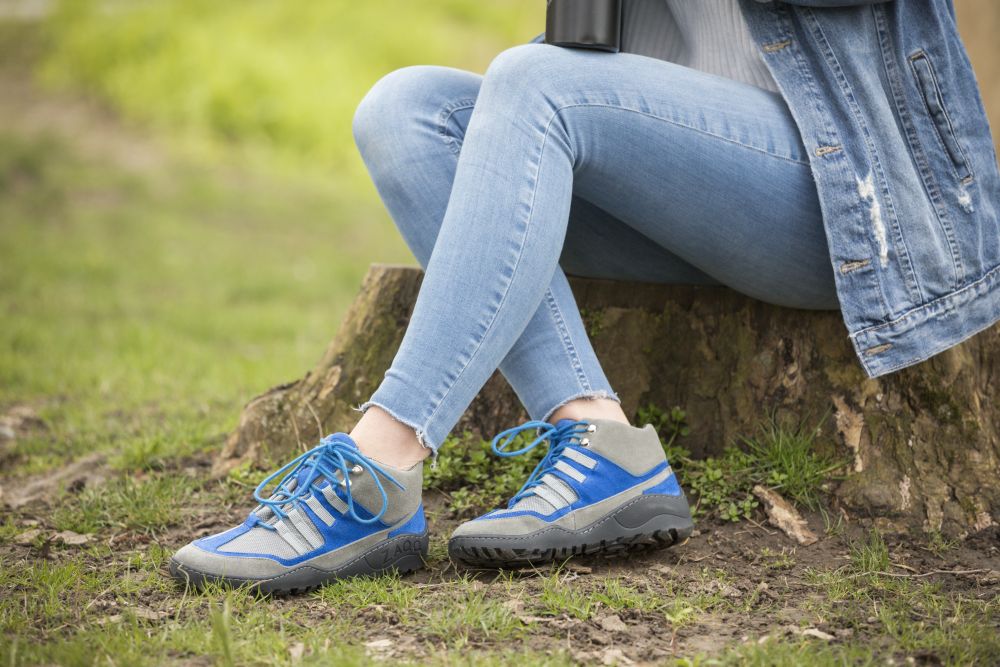 Eine Person in hellblauer Jeans, Jeansjacke und den SQOUT Grey Blue Waterproof Sneakers von ZAQQ mit blauen Schnürsenkeln sitzt auf einem Baumstumpf. Ihre Schuhe sind atmungsaktiv und fühlen sich wie Barfußschuhe an. Der Boden ist grasbewachsen und die Person hält eine Thermoskanne in der Hand.