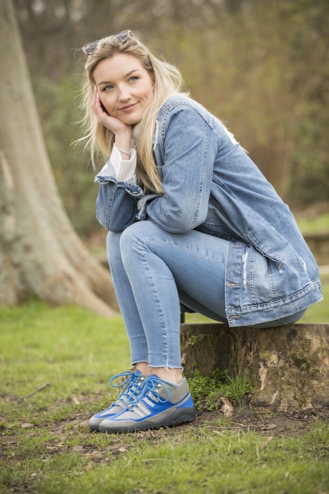 Eine Frau mit langen blonden Haaren, einer blauen Jeansjacke und Jeans bekleidet, sitzt auf einem Baumstumpf im Park. Sie ruht nachdenklich auf ihrer Hand. Ihre graublauen, wasserdichten ZAQQ SQOUT-Schuhe liegen eingebettet im Gras und verschmelzen nahtlos mit der natürlichen Umgebung der Bäume um sie herum.