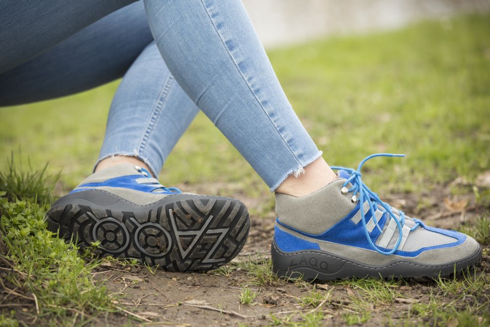 Eine Person in den für ihre atmungsaktiven Eigenschaften bekannten SQOUT Grey Blue Waterproof High-Top-Sneakern von ZAQQ sitzt mit offenen Schnürsenkeln im Gras. Nur die Beine in hellblauen Jeans sind sichtbar, während die Schuhsohlen deutliche Muster aufweisen, darunter auch der Markenname.