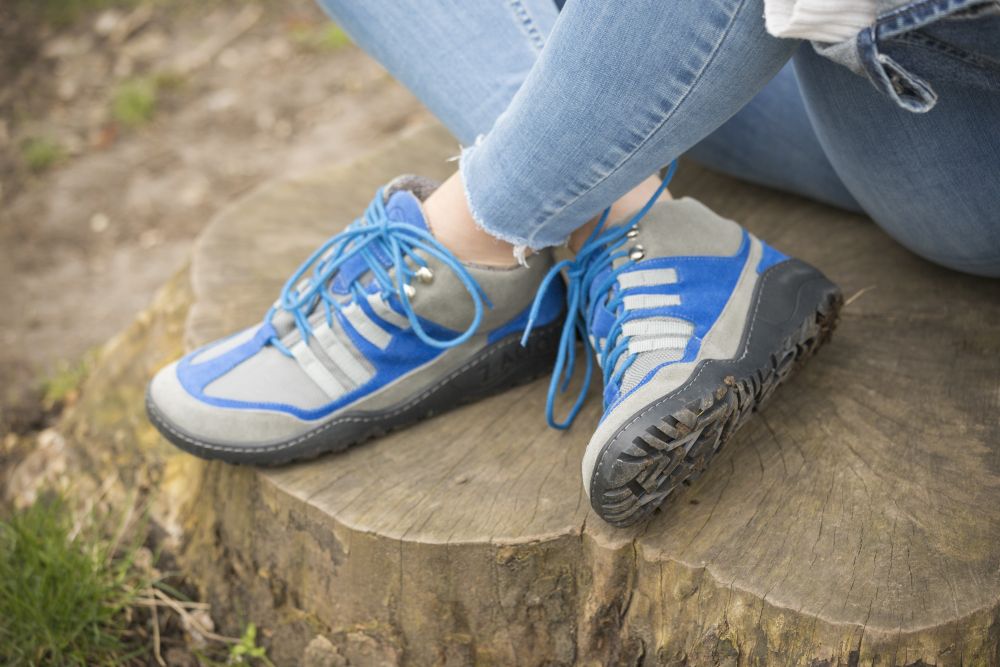 Nahaufnahme einer Person, die ZAQQs SQOUT grau-blaue wasserdichte Wanderschuhe mit blauen Schnürsenkeln trägt und auf einem Baumstumpf sitzt. Sie trägt hellblaue Jeans und der Boden um den Baumstumpf herum ist grasbedeckt und erdig.