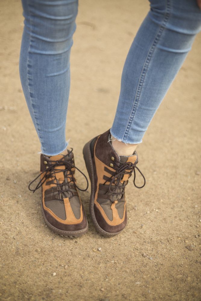 Die Beine einer Person in blauen Röhrenjeans schreiten selbstbewusst über einen sandigen Untergrund und präsentieren die braunen wasserdichten SQOUT-Schuhe von ZAQQ. Diese Stiefel verfügen über die strapazierfähige TERRA-Sohle und sind aus Veloursleder gefertigt, was Stil und Komfort bei jedem Abenteuer verspricht.