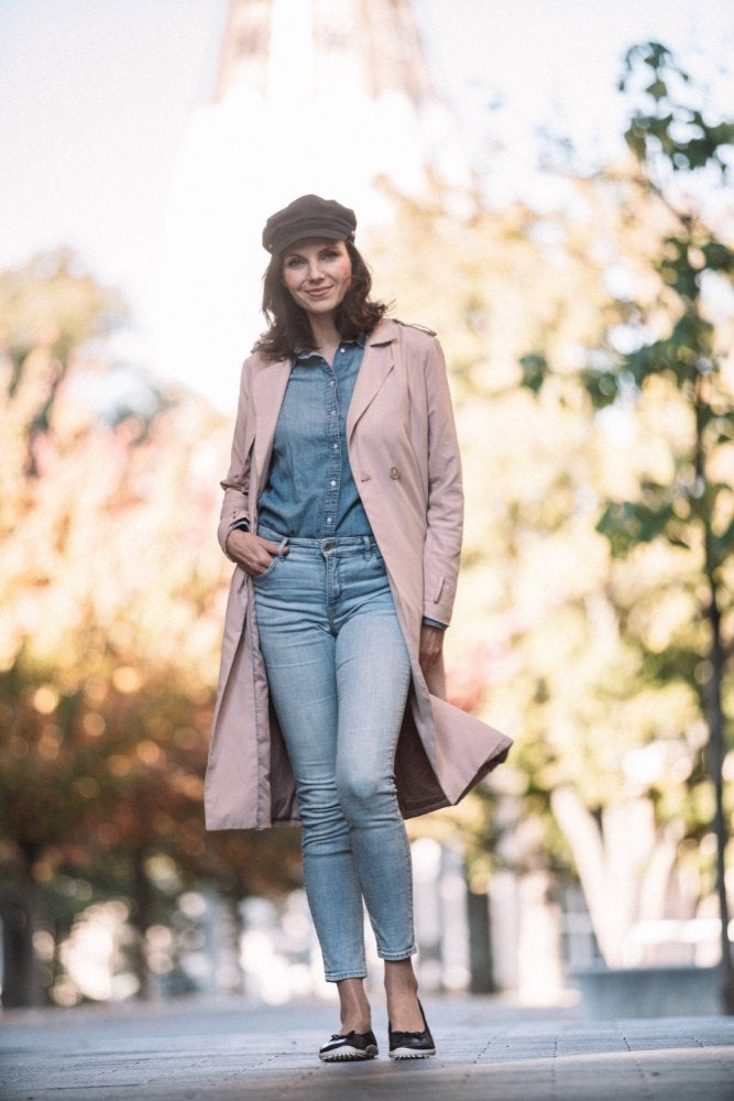 Eine Frau steht im Freien in einem hellrosa Trenchcoat, einem blauen Jeanshemd und Jeans, dazu eine schwarze Kappe. Ihre Füße sind bequem geschmückt mit schicken dunkelblauen ZAQQ SUQAR Schuhen mit Sneakersohle-Design. Mit einer Hand in der Tasche und einem Lächeln im Gesicht wird sie von Bäumen mit herbstlichem Laub im Hintergrund eingerahmt.