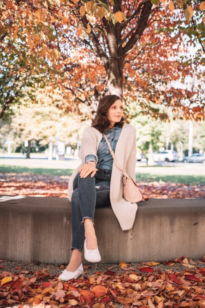 Eine Frau in einer langen beigefarbenen Strickjacke und Jeans sitzt auf einer Betonbank unter einem Baum, der mit leuchtenden Herbstblättern geschmückt ist. Sie trägt das stilvolle weiße Schuhwerk ZAQQ SUQAR, schlägt ein Bein über das andere und blickt in die Ferne, umgeben von gefallenen Blättern auf dem Boden.