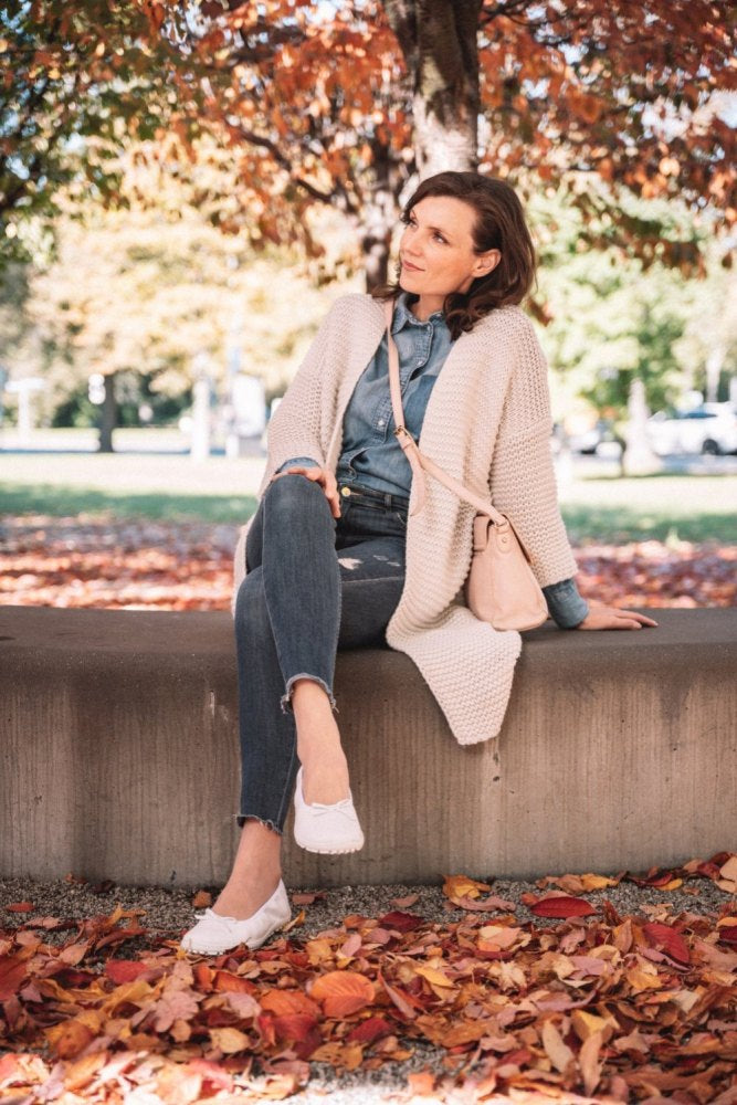 In einer herbstlichen Umgebung sitzt eine Frau in Jeanshemd, beiger Strickjacke und zerrissenen Jeans auf einer niedrigen Mauer. Ihre hellrosa Umhängetasche passt gut zu ihren weißen SUQAR-Sneakern mit Sneaker-Sohlen der Marke ZAQQ. Buntes Laub bedeckt den Boden und im Hintergrund stehen Bäume mit orangefarbenen Blättern.