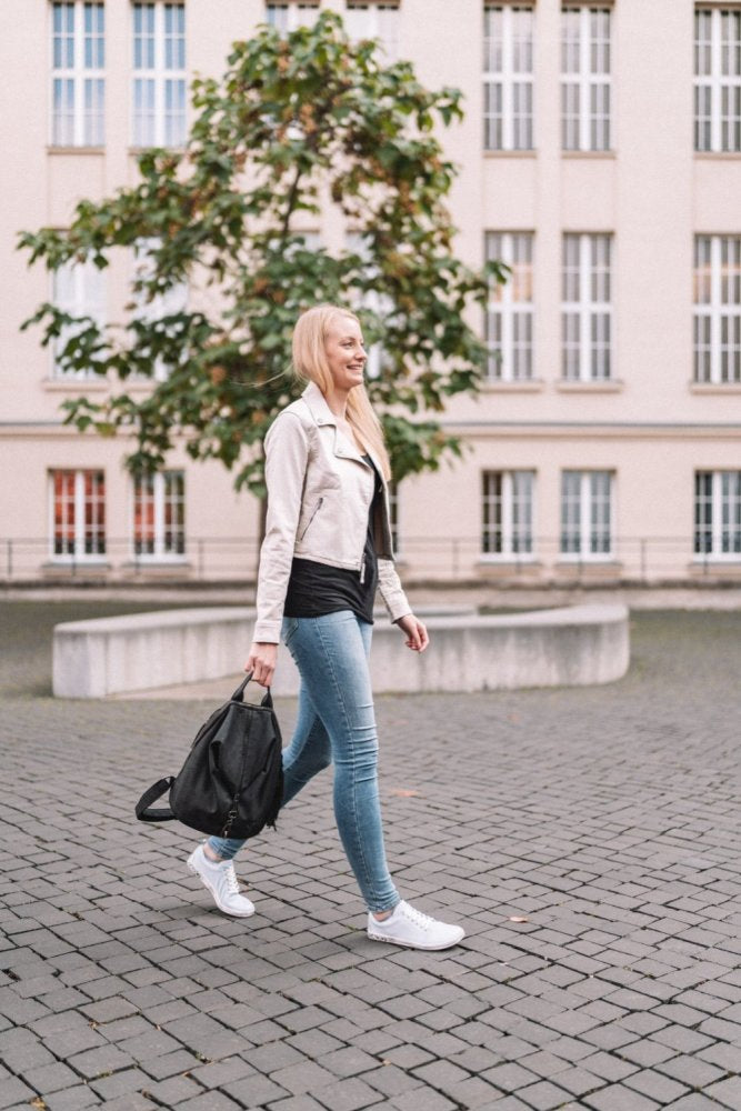 Eine Person mit langen blonden Haaren, einer hellen Jacke und Jeans läuft in ihren ZAQQ TAQQ Nappa Lightblue-Schuhen mit flexibler Sohle auf einem Kopfsteinpflasterweg. Sie trägt eine schwarze Tasche und geht an einem Innenhof mit einem großen Baum vor einem Gebäude mit hohen Fenstern vorbei.