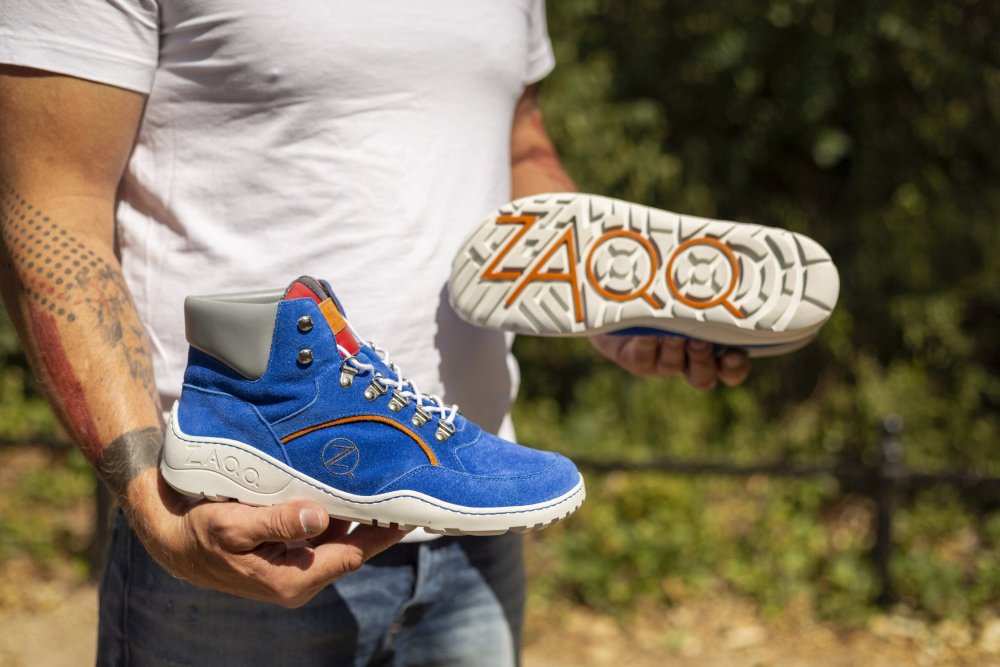 Eine Person steht im Freien und hält ein Paar blaue TERRAQ Azure Waterproof High-Top-Sneaker von ZAQQ. Ein Schuh steht aufrecht, während der andere gedreht ist, sodass die weiße Sohle mit dem ZAQQ-Logo zu sehen ist. Der lässige Look wird durch das weiße Hemd und die Jeans unterstrichen und ergibt einen entspannten Barfußschuh-Stil, der perfekt für jedes Abenteuer ist.