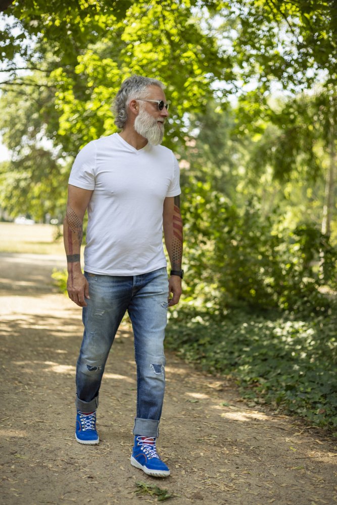 Ein Mann mit weißem Bart, weißem T-Shirt, Jeans und blauen Turnschuhen schreitet an einem sonnigen Tag einen von Bäumen gesäumten Weg entlang. Er trägt eine Sonnenbrille und Tattoos auf den Armen und strahlt bei jedem Schritt Selbstvertrauen aus, als ob er die feinsten wasserdichten TERRAQ Azure-Schuhe von ZAQQ tragen würde.