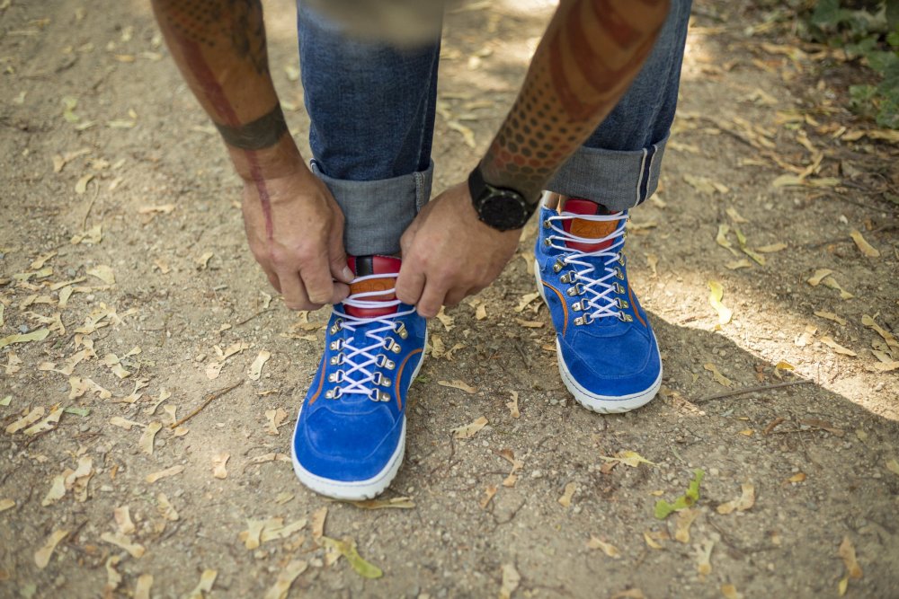 Eine Person mit tätowierten Armen schnürt die Schnürsenkel leuchtend blauer ZAQQ TERRAQ Azure Waterproof-Sneaker mit weißen und roten Details. Sie trägt hochgekrempelte Bluejeans und eine Uhr und steht auf einem Feldweg, der mit trockenem Laub übersät ist.