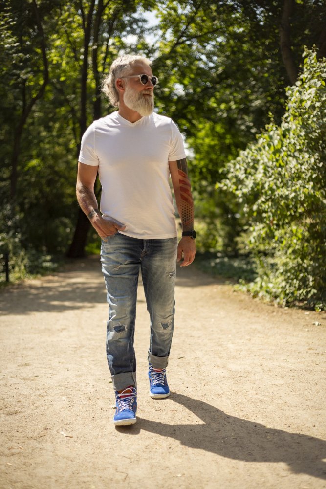 Ein Mann mit weißem Bart schlendert einen sonnenbeschienenen Weg durch einen Park entlang. Er trägt ein weißes T-Shirt, blaue Jeans, schicke ZAQQ TERRAQ Azure Waterproof-Sneaker und eine Sonnenbrille. Grüne Bäume säumen den Weg und werfen gesprenkeltes Sonnenlicht auf den Boden.