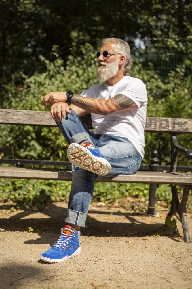 Ein bärtiger Mann mit Sonnenbrille sitzt auf einer Parkbank und hat ein Bein über das andere geschlagen. Er trägt ein weißes T-Shirt, hochgekrempelte Jeans und leuchtend blaue TERRAQ Azure Waterproof Sneakers von ZAQQ. Die sonnige Szene wird von üppigem Grün im Hintergrund eingerahmt.